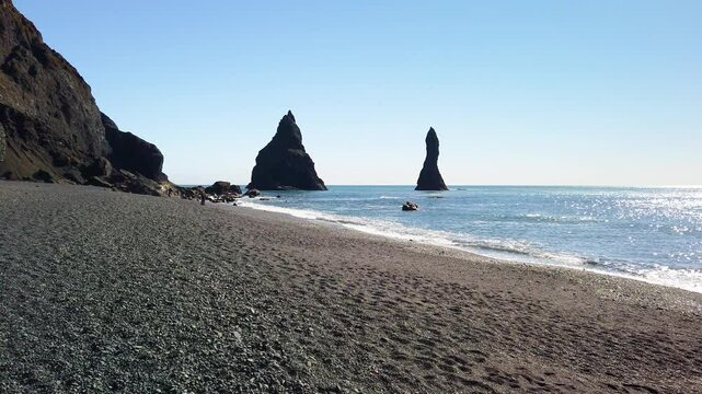 Short slow motion clip of the Reynisdrangar cliffs at the Reynisfjara black sand beach in Iceland. It is a sunny spring day, and the gentle waves from the Atlantic Ocean hits the beach