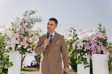 A man presenter in a beige suit leads a wedding ceremony in the park. 