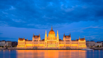 Fototapeta premium The Hungarian Parliament Building glows in warm light against a twilight sky. Reflections shimmer on the Danube River, creating a serene atmosphere in Budapest.