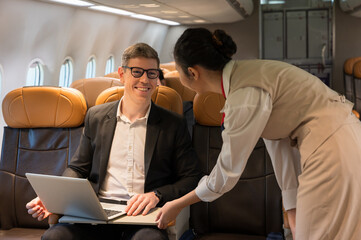 Businessman in glasses and dark suit using laptop while seat on airplane. Female flight attendant in uniform leaning over to assist smiling passenger. Work and air transportation concept
