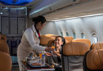 Cheerful flight attendant serving orange juice drink to smiling female passenger in airplane cabin. In flight service cart with  teapot cups water bottles and pastry