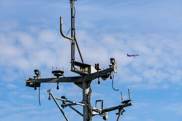 Railway catenary pole with numerous wires and technical elements, set against a blue sky with scattered clouds. In the background, a flying airplane adds an interesting contrast between ground and air