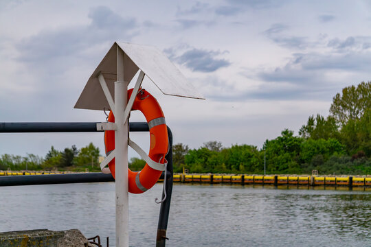 orange lifebuoy hanging on a white post with a protective roof, located at a waterfront. In the background, calm river or canal water is visible along with metal railings and lush green trees and shru