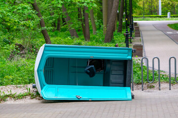 overturned portable toilet lying on a sidewalk near a park. In the background, a bicycle path,...