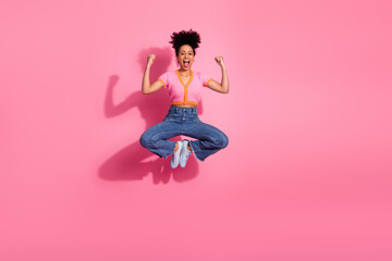 Confident young woman jumping with enjoyment in casual outfit against pink background expressing...