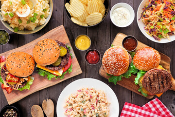 Fototapeta na wymiar BBQ table scene with hamburgers, classic salads and snacks. Top view over a dark wood background.