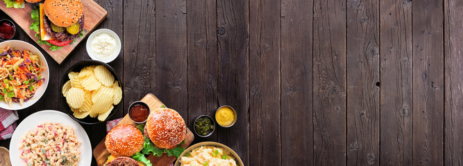 BBQ corner border with hamburgers, classic salads and snacks. Overhead view table scene over a dark wood banner background. Copy space.