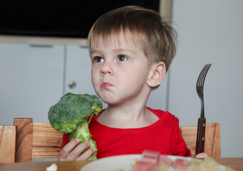 portrait of cute toddler sitting at the table with plate with noodles and sausages holding broccoli...