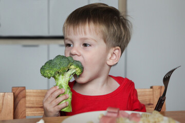 smiling toddler sitting at table with plate of noodles and sausage and holding big raw broccoli -happy mealtime at home