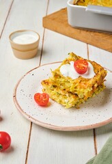 Sliced ​​cabbage, carrot and egg casserole on a plate on a light wooden background. Served with sour cream and fresh cherry tomatoes.