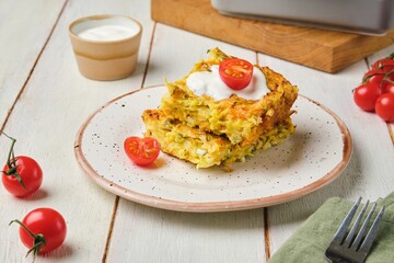 Sliced ​​cabbage, carrot and egg casserole on a plate on a light wooden background. Served with sour cream and fresh cherry tomatoes.