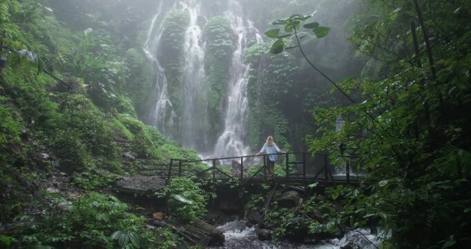 Drone shot of a blonde woman when she stands and admires the view of a waterfall in Bali jungle