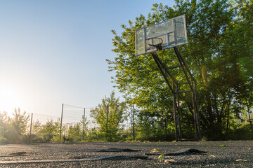 Outdoor basketball court on asphalt, visible cracks and stains, sunlight, backboard with tattered net, trees in background, green leaves, shallow depth of field