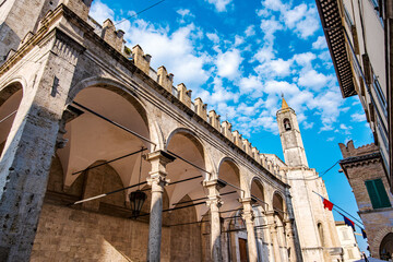 Loggia dei Mercanti featuring travertine arches and columns, framed by the bell tower of the Church of San Francesco, creates a picturesque scene under a cloudy blue sky