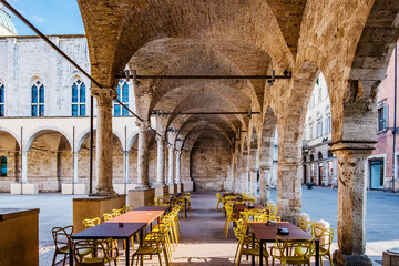 Empty tables and yellow chairs lined under brick archways in the loggia of Piazza San Francesco, Ascoli Piceno, create a serene urban architectural scene