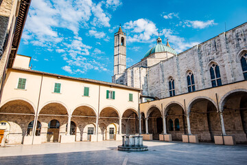 Basking in warm sunlight, the cloister of the Cathedral of Saint Emidius frames a serene courtyard alongside a majestic bell tower in Ascoli Piceno, Italy