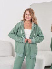 Smiling woman in green hoodie with matching pants stands confidently with hands on the jacket. She...