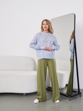 A woman in a light blue sweater and green pants stands near a floor mirror, reflecting her image. She is smiling slightly, standing on a light floor beside a white sofa
