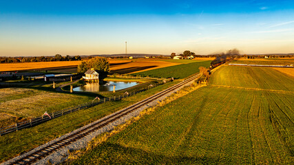 A train moves along a railroad track through expansive fields, reflecting the warm glow of the setting sun. A serene pond and quaint structures enhance the picturesque landscape.