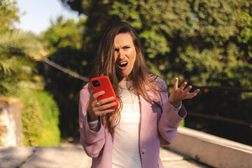 Portrait of young angry brunette long hair woman holding cellphone in hands on the street, looking at screen with irritated expression, made a mistake or annoyed by texts and calls. WTF.