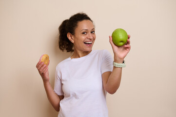 Smiling Woman Holding a Green Apple in One Hand and a Doughnut in the Other, Representing a Healthy...