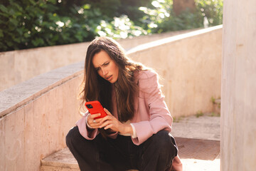 Mad and shock young brunette beautiful woman using mobile phone sitting on stairs. Unhappy woman hold mobile phone. Disappointed sad upset lady horrified impressed news. WTF. Oh no.