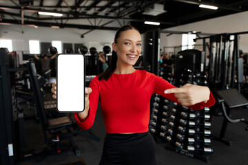Fototapeta premium Mobile mockup. Woman pointing at blank smartphone screen, indicating free space for fitness ad or website, posing in modern gym, mockup
