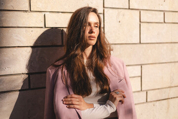 Confident young woman in a pink blazer posing with crossed arms against a modern stone wall,...