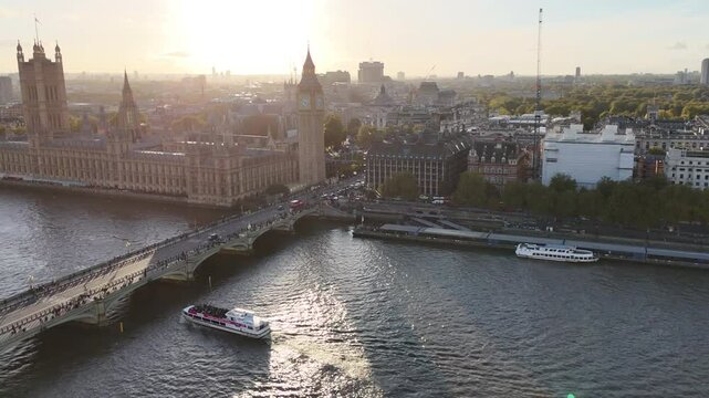 Aerial view of a bridge over the River Thames with a boat passing beneath. The London skyline unfolds in the distance, featuring Big Ben and the Westminster architecture.