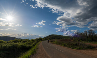 Fototapeta premium A man on a motorcycle drives on a highway that passes through hills and mountains. The sunset, white clouds and blue sky can be seen in the background. 