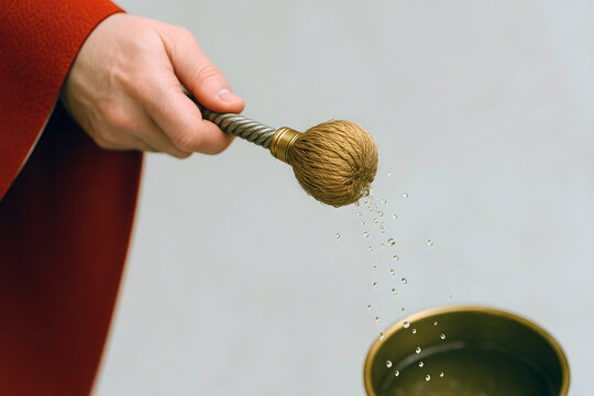 Close-up of hand sprinkling holy water with aspergillum during religious ritual, droplets in mid-air, red robe visible, clean background, symbolic Catholic purification ceremony