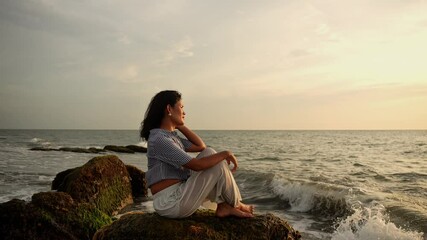 Latina woman sitting on rocks on the beach looking at the sea and the sunset in Santa Marta. High quality 4k footage - Powered by Adobe