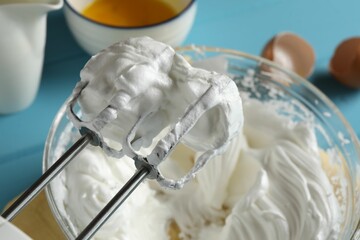 Hand mixer with whisks and whipped cream on light blue table, closeup