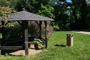 Wooden park pavillon with dark brown resin shingle roof surrounded by spring shrubs, trees and cultivated lawn. Wooden trash bin visible on the right side. 