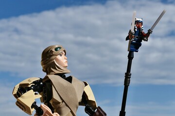 Naklejka premium LEGO Star Wars action figure of female Jedi Rey Skywalker looking at small figure of female Togruta Jedi Ahsoka Tano with two lightsabers on tip of her pole. Cloudy skies background. 