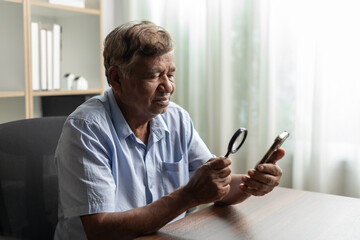Senior man with vision problems using magnifying glass to look at text on smartphone while sitting at his desk at home.