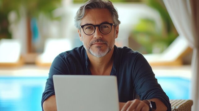 middle-aged man with gray hair and beard wearing glasses working on laptop by a poolside with a relaxed and focused expression