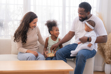Fototapeta premium Happy family sitting together on a sofa, spending quality time and smiling while enjoying snacks at home in a warm and cozy living room.