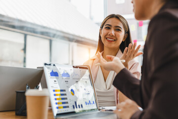 Businesswoman and colleague discussing data analysis with charts on laptop in modern office, smiling and engaged in teamwork, coffee cup on table, bright daylight atmosphere