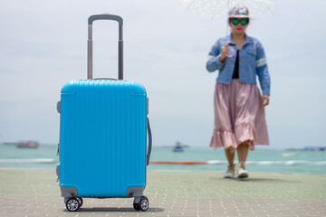 Bright Blue Luggage on a Pier with a Traveler and Sea View. Vibrant blue hard-shell suitcase stands prominently on a seaside pier, with a person holding an umbrella walking in the blurred background