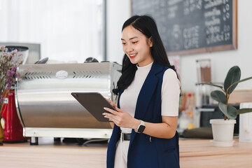 Young Asian woman in smart casual outfit stands in modern coffee shop, smiling while using digital tablet, surrounded by coffee equipment and cozy decor