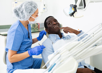 Obraz premium African-american man pointing at his teeth during consultation, woman dentist sitting and listening to him.