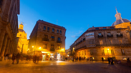 Obraz premium Scenic view of busy Plaza de Anaya in Spanish city of Salamanca overlooking ancient illuminated buildings, domes of Clerecia and Palace de Anaya against blue sky on spring evening