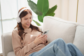 Asian Young woman listening to the music with white headphones smiles while using her smartphone on a couch