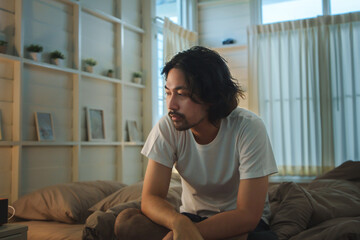 Asian man with medium-length hair sitting on bed in morning light wearing a plain white T-shirt,...