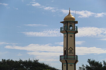 A tall mosque minaret with a golden dome under a bright blue sky, standing out against trees and...