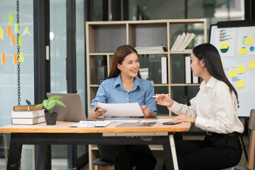Fototapeta premium Two Asian businesswomen holding graph papers are sitting and talking happily while working. 