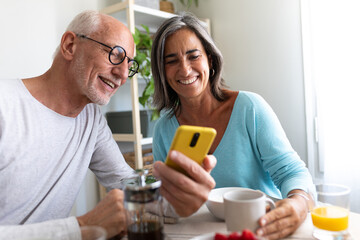 Happy mature caucasian married couple enjoying healthy breakfast at home looking at phone.