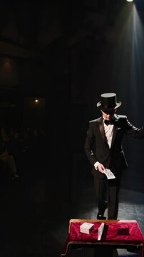 A magician in a tuxedo and top hat performs under a spotlight on stage. Low-angle shot creates a dramatic effect, ideal for a magic show video.