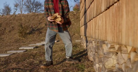 Guy in a hat and a plaid shirt collects firewood in an armful while relaxing in his country house. Guy collects firewood while relaxing in a country house